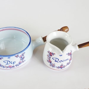 Op de afbeelding: Twee keramische serveerschalen. Eén is een ondiepe kom met een handvat, het woord "Dip" en bloemaccenten. De andere is een kleine kan met een houten handvat en het woord "Room". Beide hebben blauwe en roze details.