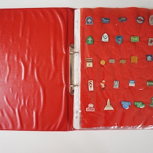 Op de afbeelding: Een rode map met een transparant plastic vel erin. Het vel bevat een verzameling vintage emaille-pins met verschillende logo's en ontwerpen.