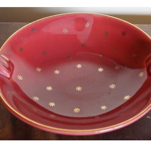 Op de afbeelding: Een rode keramische serveerschaal met gouden sterrenpatronen en gouden rand. De schaal heeft twee geribbelde handvatten en een glanzende afwerking, geschikt voor het serveren van voedsel of als decoratief stuk.