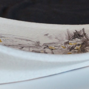 May include: A white ceramic dish with a curved shape and a stylized landscape design in brown and yellow paint.