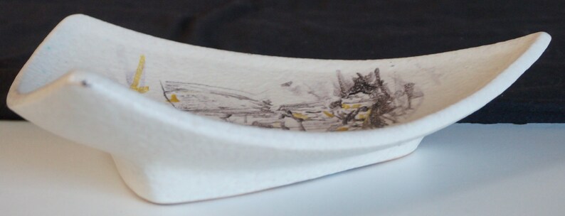 May include: A white ceramic dish with a curved shape and a stylized landscape design in brown and yellow paint.