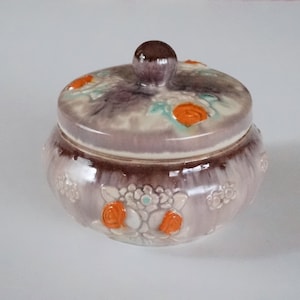 以下が含まれることがあります： 紫、オレンジ、白の色の花柄で飾られた蓋付きの陶器の壺。壺は光沢のある仕上げで、丸みを帯びた形をしています。