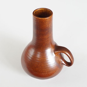 May include: A brown ceramic vase with a rounded body and a cylindrical neck. The vase has a small handle on the side. The glaze has a glossy finish. The vase is a decorative item.