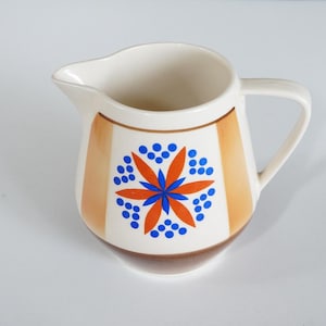 May include: Cream ceramic creamer with a brown base and handle. It has vertical brown stripes and a central floral design in orange, blue, and white. A classic kitchenware item.