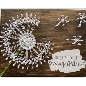 Könnte beinhalten: DIY String Art Kit mit einem Pusteblumen-Design. Weißer Faden bildet die Blume und die Samenköpfe auf einer dunkelbraunen Holzplatte. Der Text "DO IT YOURSELF String Art Kits" ist sichtbar.