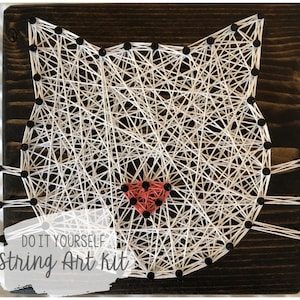 Puede incluir: Kit de arte de hilo para hacer tú mismo con un rostro de gato blanco con una nariz roja sobre un fondo de madera oscura. El texto "DO IT YOURSELF String Art Kit" está impreso en la parte inferior de la madera.
