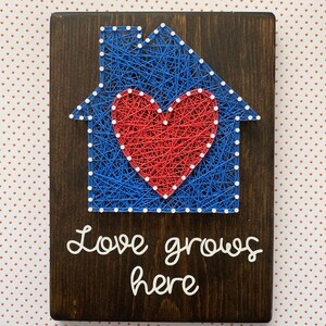 May include: A wooden sign with a blue and red string art design of a house with a heart inside. The text "Love grows here" is written below the house.