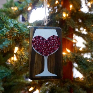 May include: A wooden ornament shaped like a wine glass with a red heart made of string art inside. The ornament has a rustic brown finish and a white background.