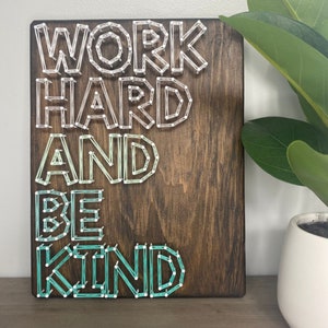 May include: A wooden sign with the phrase "Work Hard and Be Kind" spelled out in colourful string art. The letters are made with white, light blue and green string.