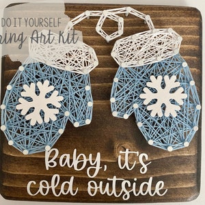 May include: DIY string art kit featuring two blue and white mittens with snowflakes on a brown wooden background. The text "Do It Yourself String Art Kit" and "Baby, it's cold outside" is written in white on the wood.