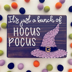 May include: A wooden sign with the text "It's just a bunch of HOCUS POCUS" in white lettering. A purple witch's hat with a black band and gold buckle is string art on the sign.