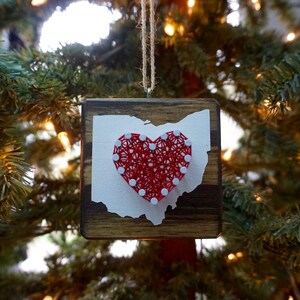 Puede incluir: Un adorno de madera con forma del estado de Ohio con un corazón de cuerda roja en el centro. El corazón está hecho con cuerda blanca envuelta alrededor de clavos.