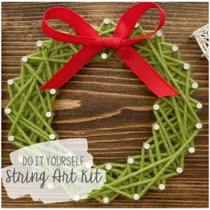 May include: Green yarn is wrapped around nails to create a wreath shape. A red ribbon with a bow is at the top of the wreath. The text "DO IT YOURSELF string Art kit" is written on a white background.