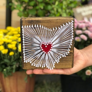 May include: A wooden plaque with a red heart in the center of an outline of the state of Ohio. The outline is created with white string wrapped around nails hammered into the wood.