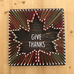 May include: A wooden sign with a string art design of a leaf in shades of orange, red, and green. The sign says "Give Thanks" in white lettering.