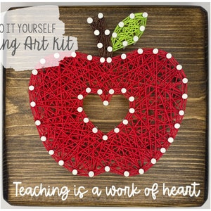 May include: DIY string art kit featuring a red apple with a heart shape inside. The wooden plaque has the text "Teaching is a work of heart".