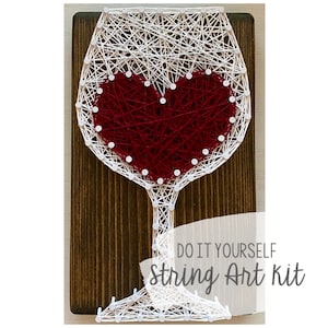 Puede incluir: Arte de hilo blanco de una copa de vino con un corazón rojo en su interior. El arte está sobre un fondo de madera con el texto "Do It Yourself String Art Kit".