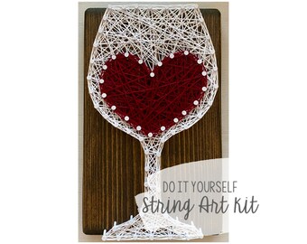 May include: White string art of a wine glass with a red heart inside. The art is on a wooden background with the text "Do It Yourself String Art Kit".