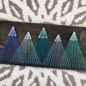 Könnte beinhalten: Ein String-Art-Werk, das eine Bergkette mit einem Farbverlauf aus blauem, grünem und weißem Garn darstellt. Die Berge werden durch das Umwickeln von Garn um Nägel geschaffen, die in eine dunkle Holzplatte geschlagen wurden.