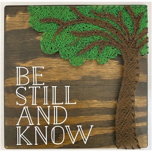 May include: A wooden sign with a green and brown string art tree design. The sign says "Be Still and Know" in white lettering.