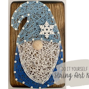 May include: DIY string art kit featuring a blue and white gnome with a snowflake on its hat. The kit includes all the materials needed to create the artwork, including string, nails, and a wooden base.