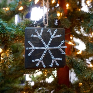 May include: A white string snowflake ornament on a dark brown wooden background. The snowflake is made with white string wrapped around nails hammered into the wood.