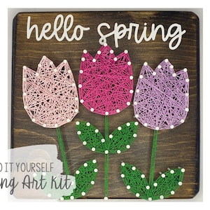 Könnte beinhalten: DIY-String-Art-Set mit drei rosa, lila und grünen Tulpen mit weißen Punkten auf einem Holzgrund. Der Text "hello spring" ist über den Tulpen geschrieben.