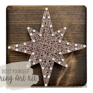 Op de afbeelding: DIY string art-set met een roségouden ster op een bruine houten basis. De set bevat alle materialen die nodig zijn om de ster te maken, inclusief touw, spijkers en een houten basis.