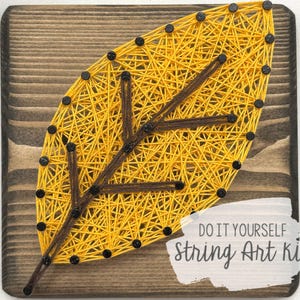 Puede incluir: Un kit de arte de cuerda de bricolaje con un diseño de hoja amarilla sobre un fondo de madera. El kit incluye todos los materiales necesarios para crear la obra de arte, incluyendo cuerda, clavos y una tabla de madera. El texto "DO IT YOURSELF string Art kit" está impreso en la tabla.