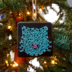 Könnte beinhalten: Ein tealblauer Katzenkopf aus String Art auf einem dunklen Holzgrund. Die Nase der Katze ist rosa und die Augen sind schwarz.