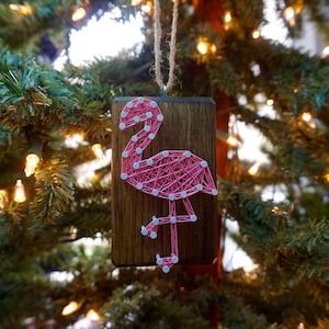 May include: Pink flamingo string art ornament on a wooden plaque. The flamingo is made with pink string wrapped around nails. The nails are white and the background is brown.