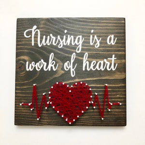 May include: Wooden sign with the text "Nursing is a work of heart" in white lettering. A red heart with white dots is strung with red thread, and a white thread EKG line is strung below the heart.