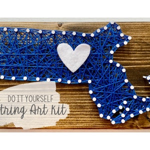 Peut inclure: Kit de broderie en fil pour réaliser soi-même un tableau représentant la silhouette du Massachusetts avec un cœur en feutrine blanche au centre. La broderie est réalisée sur une base en bois avec le texte "DO IT YOURSELF string Art kit" écrit en dessous.