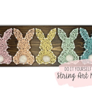 Op de afbeelding: Een DIY string art-set met vijf pastelkleurige konijnen op een houten plank. De tekst "DO IT YOURSELF String Art Kit" is gedrukt op een roze achtergrond.