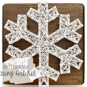 Può includere: Kit fai da te per string art con un design a fiocco di neve. Filo bianco teso tra chiodi su una tavola di legno marrone scuro. Il testo "DO IT YOURSELF String Art Kit" è visibile. Ideale per decorazioni invernali.