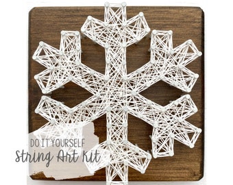 Peut inclure: Kit de bricolage de string art avec un motif de flocon de neige. Du fil blanc est tendu entre des clous sur une planche de bois brun foncé. Le texte "DO IT YOURSELF String Art Kit" est visible. Idéal pour la décoration hivernale.