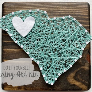 May include: DIY string art kit featuring a teal outline of South Carolina with a white felt heart in the center. The art is mounted on a dark wood background.