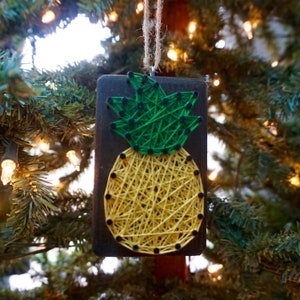 Op de afbeelding: Een houten ananas ornament met een geel en groen string art ontwerp. De ornament hangt aan een kerstboom.