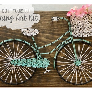 Puede incluir: Kit de arte de hilo DIY con una bicicleta con una cesta de flores. La bicicleta está hecha de hilo blanco y turquesa envuelto alrededor de clavos negros en una tabla de madera.