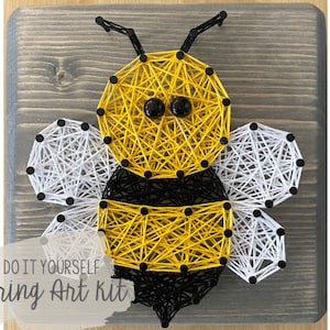 May include: DIY string art kit featuring a bee with yellow and black stripes, white wings, and black eyes. The bee is strung on a wooden board with black tacks.
