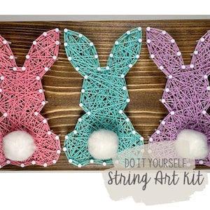 Op de afbeelding: Drie string art konijnensets op een houten achtergrond. De konijnen zijn roze, turquoise en paars. De sets bevatten alle materialen die nodig zijn om de string art te maken.