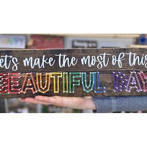 May include: Wooden sign with the text "Let's make the most of this beautiful day" in white lettering. The word "beautiful" is spelled out in rainbow colored yarn.