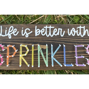 Può includere: Un cartello rettangolare in legno marrone scuro con la frase "Life is better with SPRINKLES" in bianco e colori arcobaleno. La parola "SPRINKLES" è realizzata con arte di filo colorato, aggiungendo un tocco giocoso.