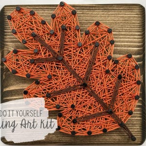 Puede incluir: Kit de arte de hilo para hacer tú mismo con un diseño de hoja naranja sobre un fondo de madera. El texto "Do It Yourself String Art Kit" está impreso debajo de la hoja.