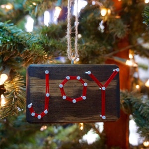 以下が含まれることがあります： 赤い糸が銀色の釘に巻き付けられ、木製のクリスマスオーナメントに「喜び」の文字が書かれています。