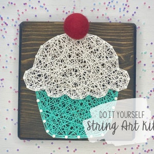 Può includere: Kit di arte con filo fai-da-te con un cupcake con una ciliegia rossa in cima. Il cupcake è fatto di filo bianco e turchese avvolto attorno a chiodi su una tavola di legno. Il testo "DO IT YOURSELF String Art Kit" è stampato sulla tavola.