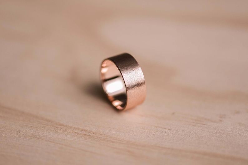 May include: A brushed rose gold wedding band with a wide, flat profile.