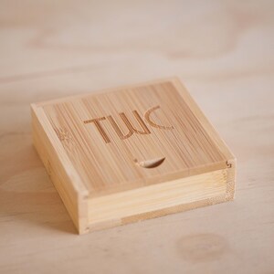 May include: A small, square, wooden box with the word "TWIC" engraved on the top. The box is made of light brown bamboo.
