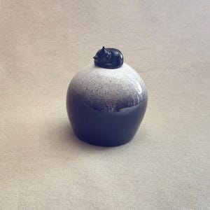Op de afbeelding: Een decoratieve keramische pot met een gespikkelde wit-grijze bovenkant en een donkerblauwe basis. Bovenop staat een kleine, glanzend zwarte kattenfiguur. De pot heeft een afgeronde vorm en een gladde, glanzende afwerking. Ongeveer 10 cm hoog.