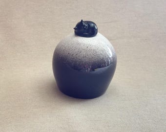 Puede incluir: Un jarrón decorativo de cerámica con una parte superior moteada en blanco y negro y una base azul oscuro. Una pequeña figura de gato negro brillante descansa en la parte superior. El jarrón tiene forma redondeada y un acabado liso y brillante. Aproximadamente 10 cm de altura.